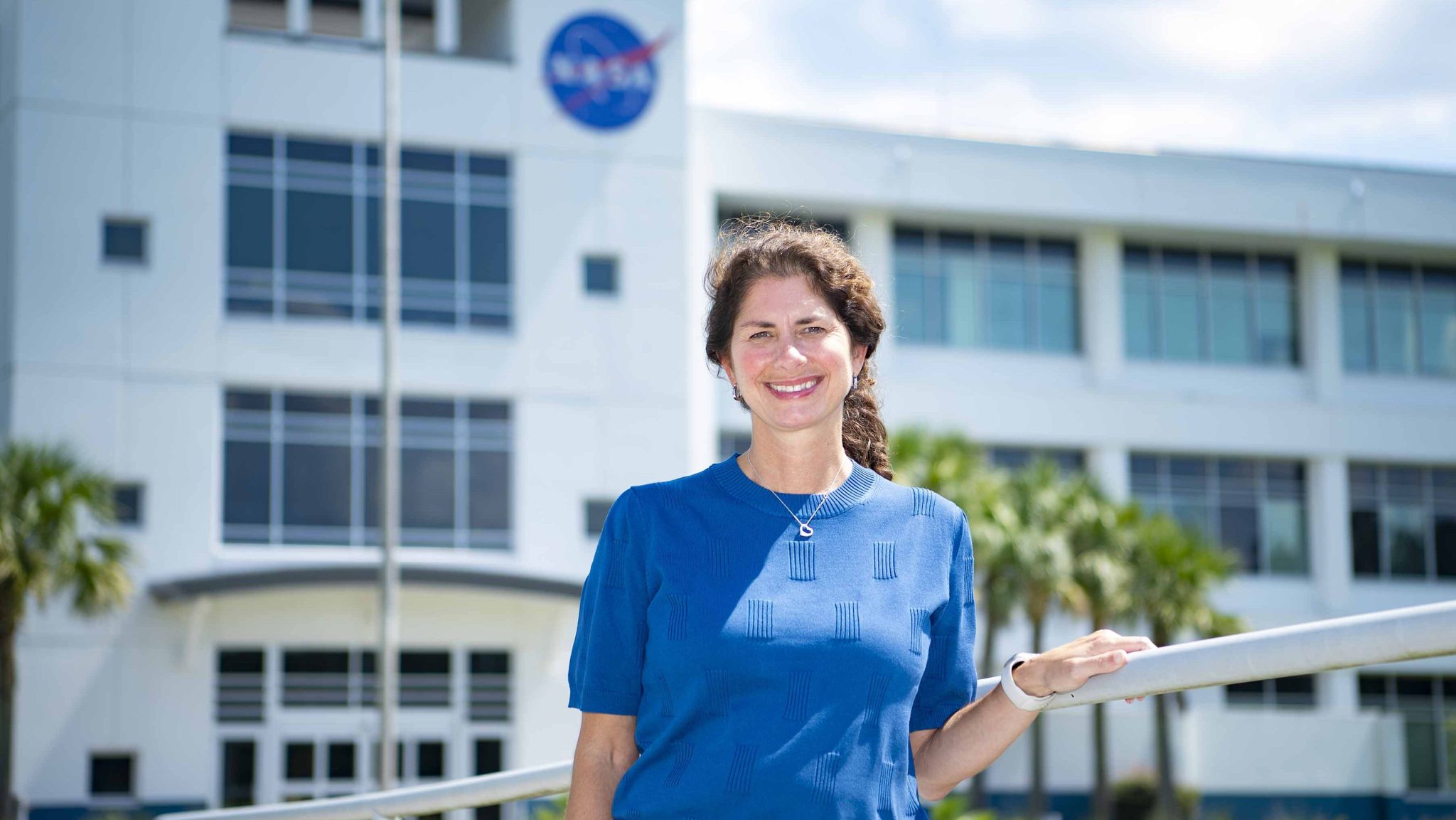 Bridget Moody, wearing a blue sweater, smiles for a portrait in front of Building 1111 at Stennis Space Center