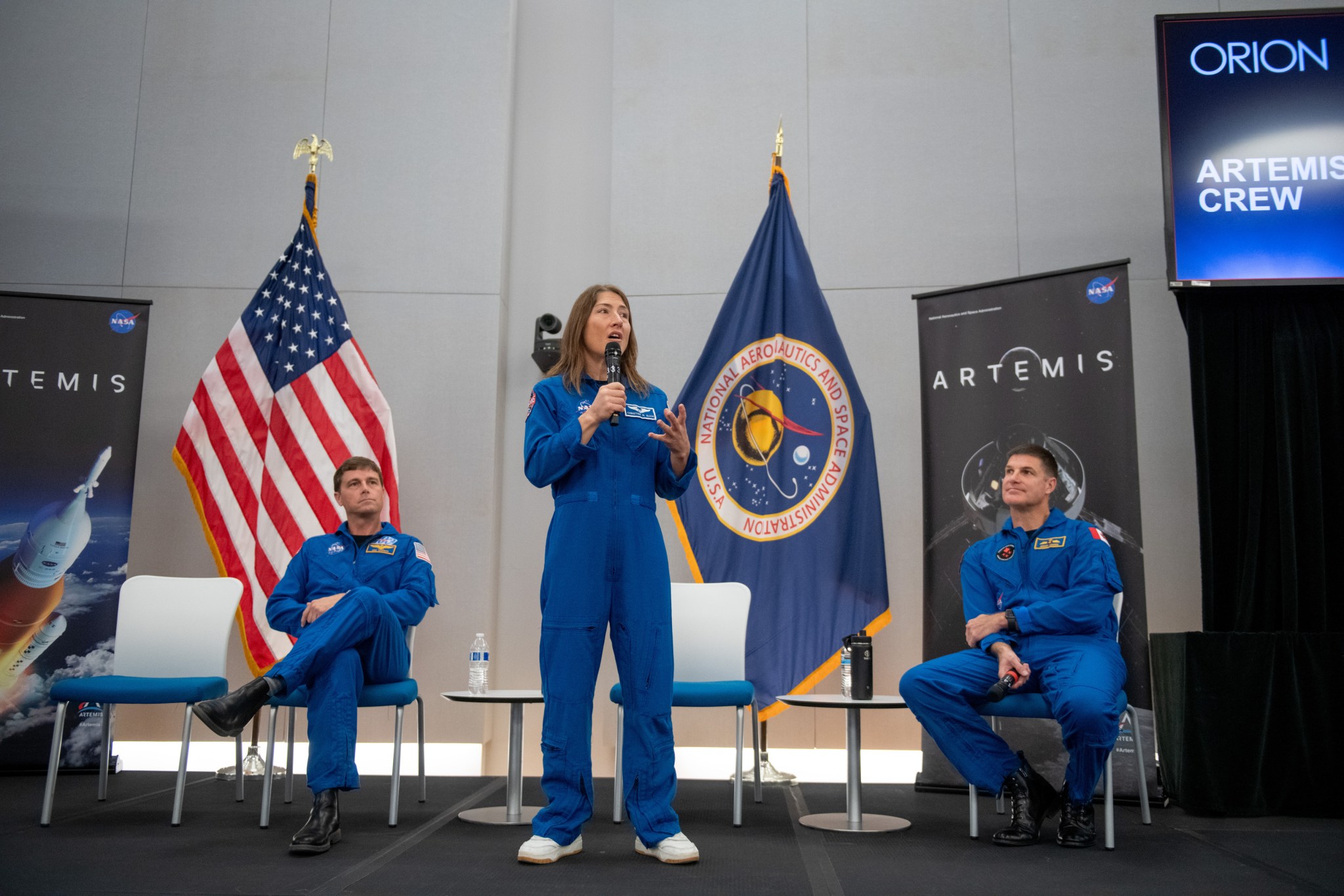 Two male astronauts are seated on either side of a female astronaut who is standing and talking into a microphone. They are dressed in blue flight suits and are on a stage. An American flag and a NASA flag, along with banners stating “Artemis,” are in the background.