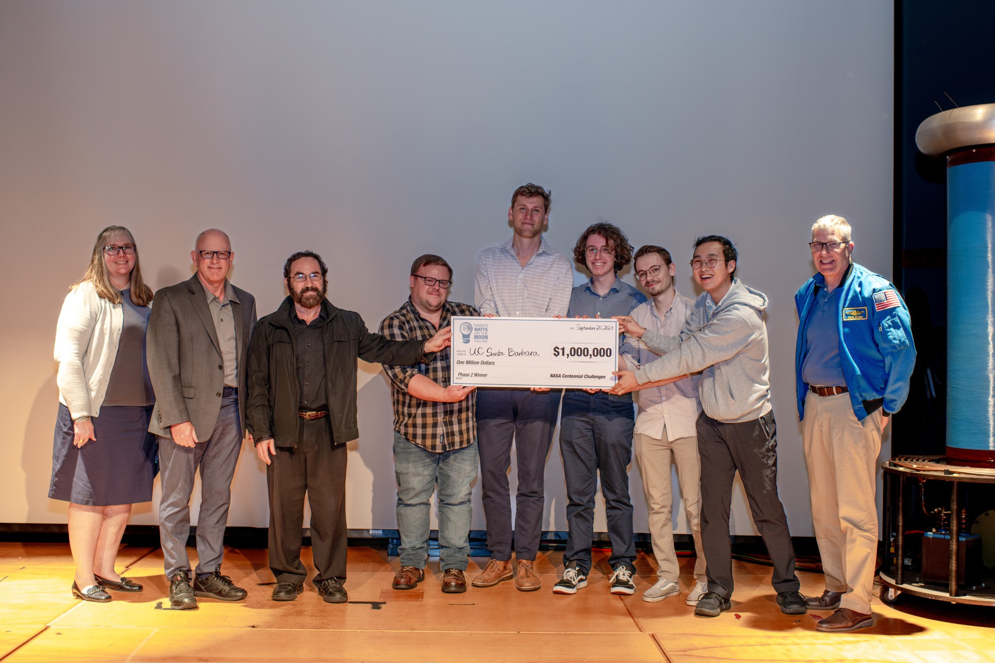 Nine people stand on stage with several of them holding parts of a large check for $1 million.