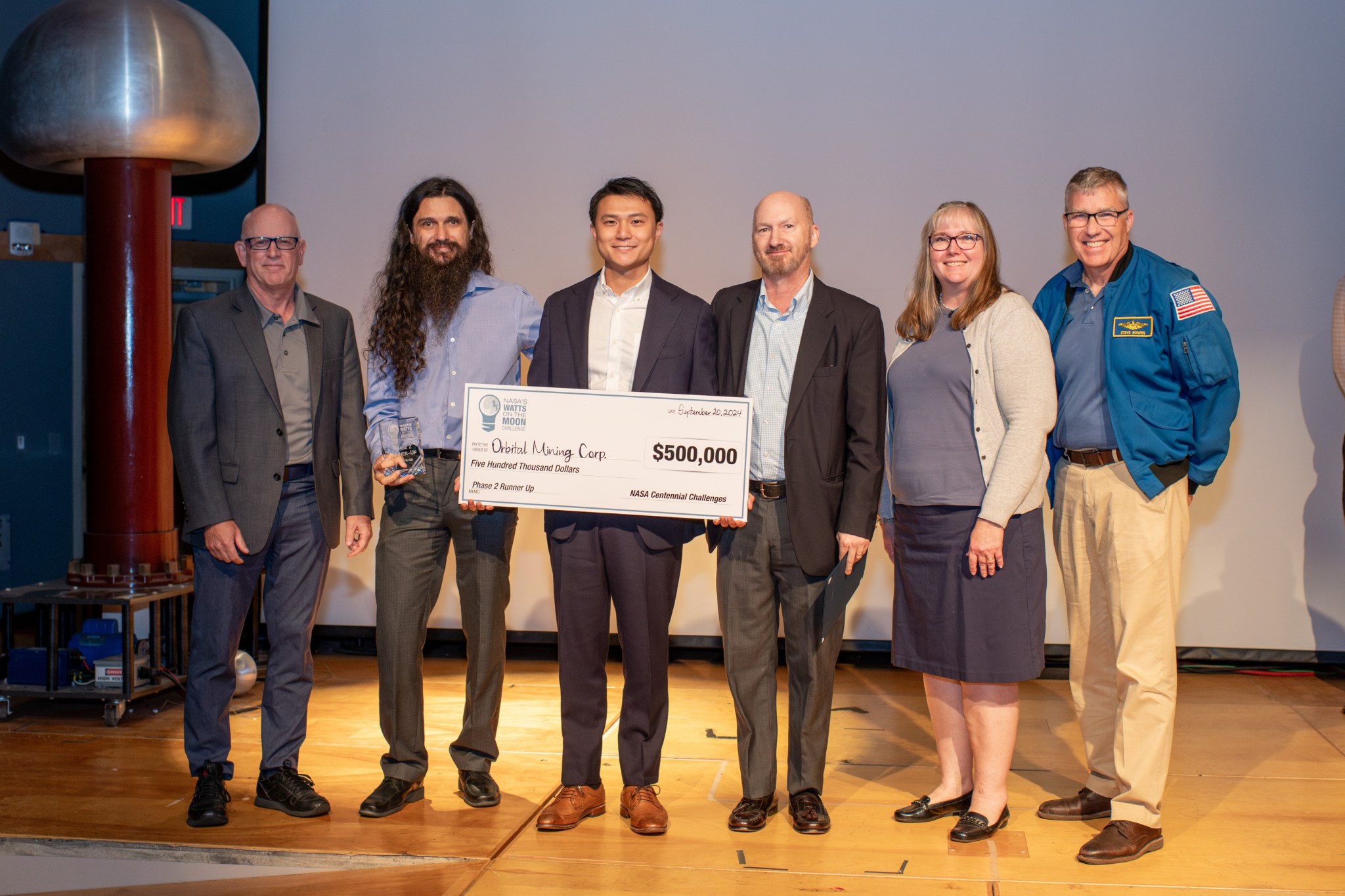 Six people stand on stage with the person in the center holding a large check for $500,000.