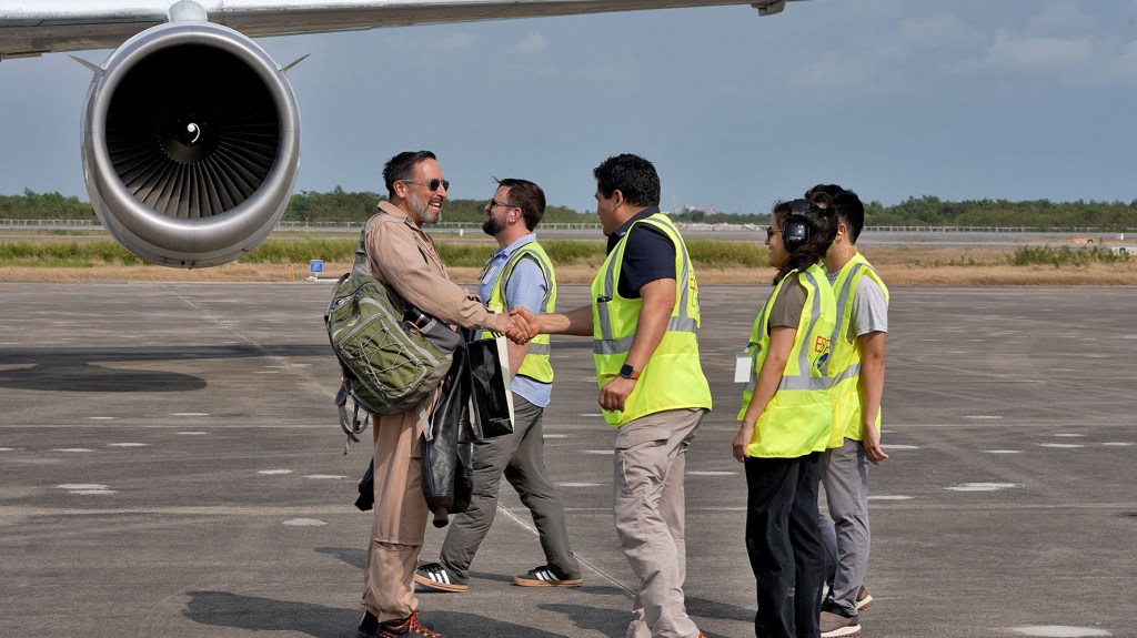 Four people in neon yellow vests welcome a man in a tan flight suit with a green backpack, standing on a gray tarmac. In the background, top left of the screen is an airplane engine.