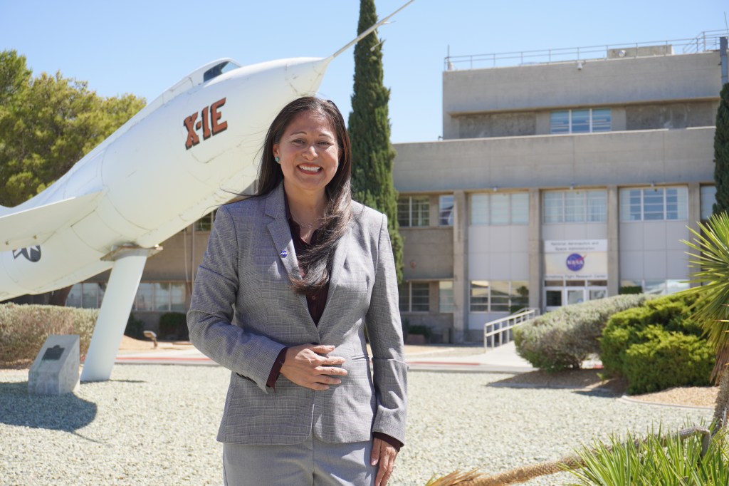 A woman in a gray suit stands smiling in front of a NASA building. Behind her to the left, the X-1E experimental aircraft is displayed.