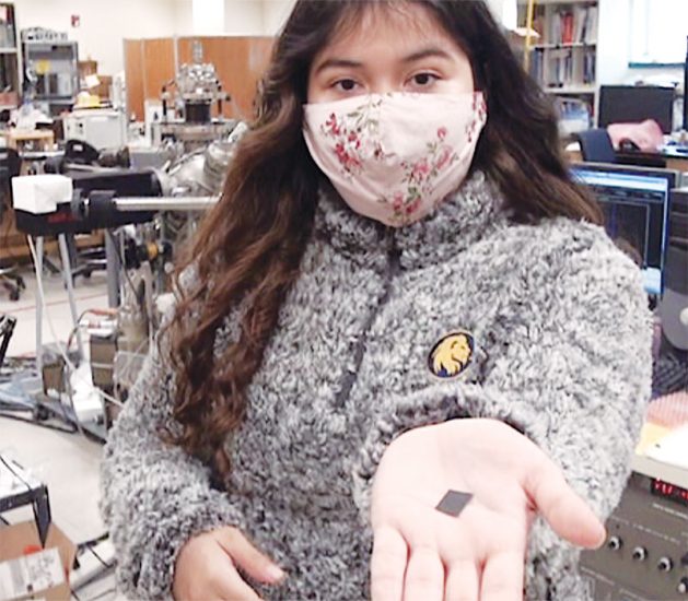 A person wearing a floral face mask and a fluffy grey sweater is holding a small, square object in their hand. They are standing in an equipment-filled laboratory or workshop space.