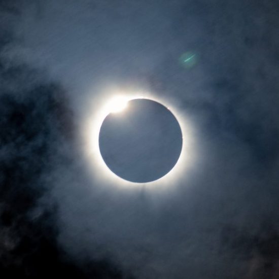 A solar eclipse is seen with the moon almost completely covering the sun. A bright ring of sunlight forms a halo around the black silhouette of the moon, with a small part of the sun peeking out from the top left. Dark clouds partially obscure the view.